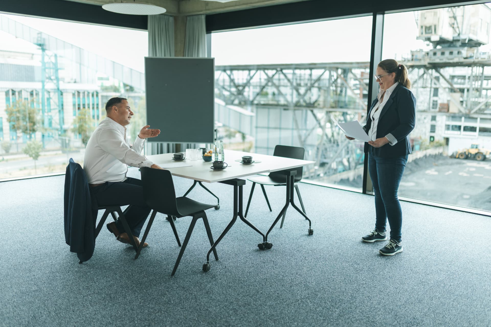 Aufnahme aus einem Besprechungstermin: Eine Frau mit Dokumenten steht am Tisch, ein Mann sitzt ihr gegenüber und erläutert etwas.